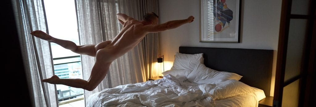 male escort jumping onto a hotel bed in a superman pose